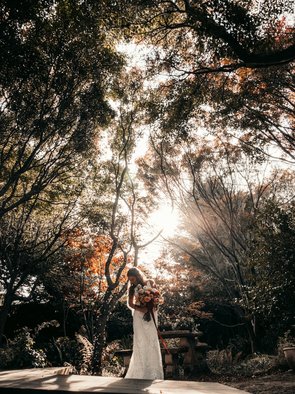 Frau in weißem Kleid tagsüber auf brauner Holzbank unter braunen Bäumen