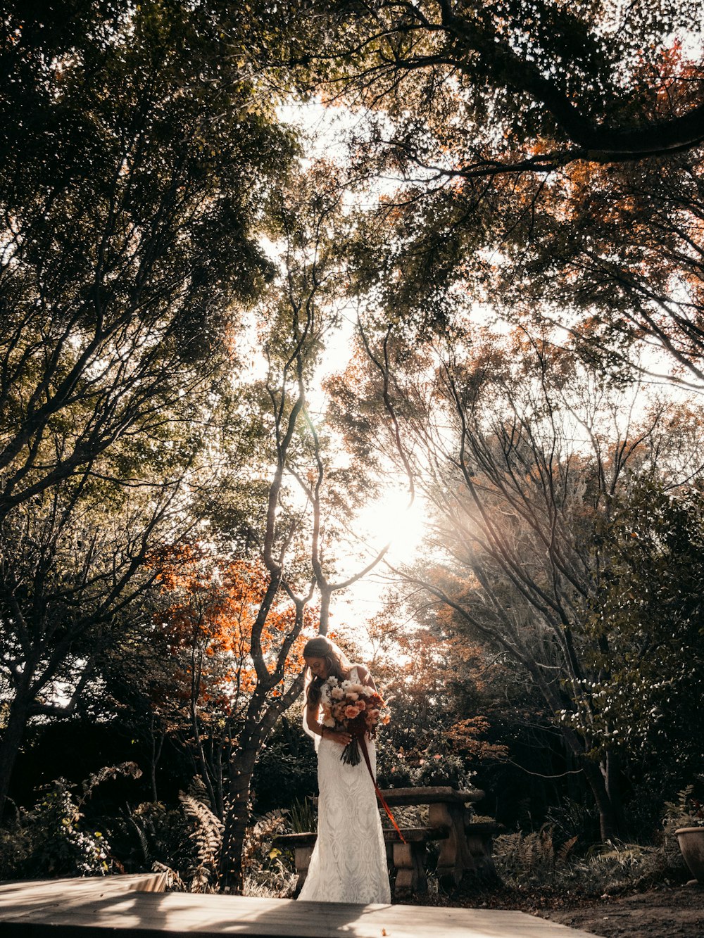 woman in white dress standing on brown wooden bench under brown trees during daytime