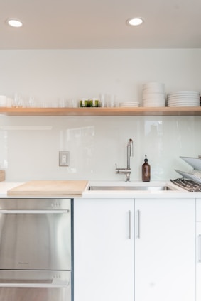 A modern minimalist kitchen setup with white cabinets and sleek stainless steel fixtures. A wooden shelf above holds stacked white plates and glass cups. A stainless steel sink is accompanied by a contemporary faucet and a brown bottle, likely for soap. A cutting board is positioned near the sink, and a dish rack is placed on the right side.