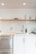 A modern minimalist kitchen setup with white cabinets and sleek stainless steel fixtures. A wooden shelf above holds stacked white plates and glass cups. A stainless steel sink is accompanied by a contemporary faucet and a brown bottle, likely for soap. A cutting board is positioned near the sink, and a dish rack is placed on the right side.