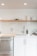 A modern minimalist kitchen setup with white cabinets and sleek stainless steel fixtures. A wooden shelf above holds stacked white plates and glass cups. A stainless steel sink is accompanied by a contemporary faucet and a brown bottle, likely for soap. A cutting board is positioned near the sink, and a dish rack is placed on the right side.