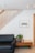 A modern living room wall with wooden vertical slats running diagonally, complemented by a black sofa on the left. A small wooden side table holds a potted plant and books. A framed artwork of birds is on the wall above the sofa.