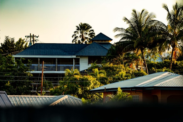A tropical setting featuring a two-story house with a dark roof surrounded by lush greenery and palm trees. The scene is bathed in warm, golden light, possibly during sunset or sunrise, creating a serene and picturesque atmosphere. Power lines are visible in the foreground, adding a hint of urban elements to the otherwise natural landscape.