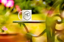 A white coffee mug featuring the logo 'HD Media' is placed on a green outdoor table, surrounded by vibrant and blurred pink and green foliage.