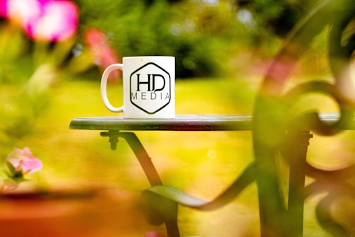 A white coffee mug featuring the logo 'HD Media' is placed on a green outdoor table, surrounded by vibrant and blurred pink and green foliage.