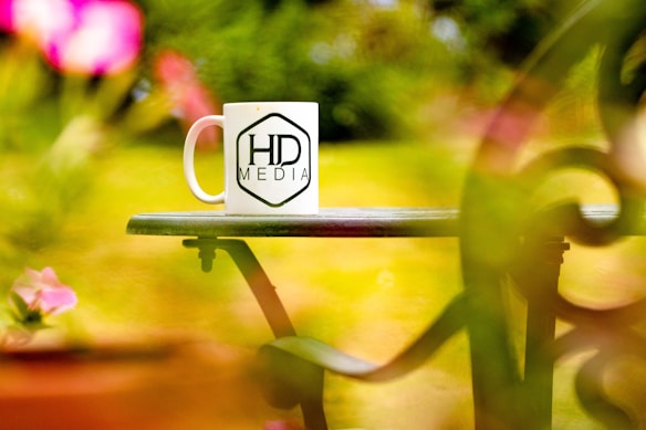 A white coffee mug featuring the logo 'HD Media' is placed on a green outdoor table, surrounded by vibrant and blurred pink and green foliage.