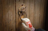 A hand wearing a knitted glove with a red and white pattern is holding a rustic, ornately designed metal door knocker fixed onto a wooden door. The glove has a winter theme, and the door has a weathered appearance.