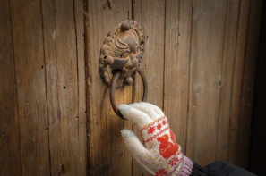 A hand wearing a knitted glove with a red and white pattern is holding a rustic, ornately designed metal door knocker fixed onto a wooden door. The glove has a winter theme, and the door has a weathered appearance.