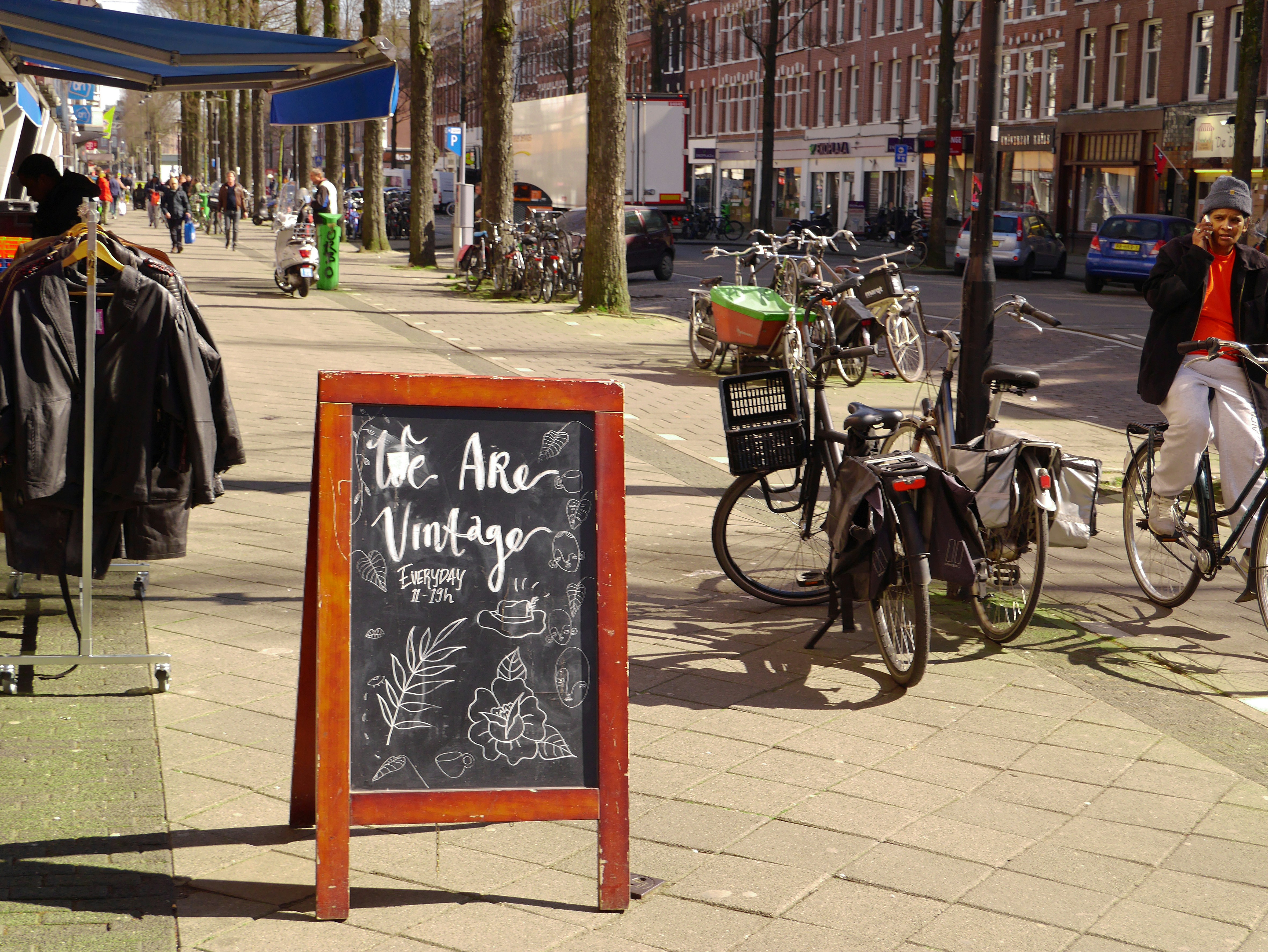 black city bike parked beside green bicycle during daytime, Free photo of a shopping street with people in Amsterdam city - in the district Dapperbuurt. It is in the light of early Spring. Picture of 26 March 2020; urban photography of The Netherlands. In Dutch: foto van van de winkelstraat, Eerste van Swindenstraat in de Dapperbuurt van Amsterdam. 26 maart 2020 - stadsfotografie, Fons Heijnsbroek.