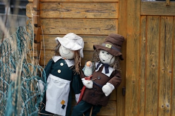 Two ragdoll figures with cheerful expressions are propped against a rustic wooden wall. The dolls are wearing vintage-style clothing; one is in a dark green dress with a white apron and cap, while the other is dressed in a brown suit with a matching hat. A small plush bird rests on one of their arms. The background has a wooden texture and there is a green netting visible on the left side.