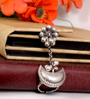 A stack of books is adorned with decorative covers, featuring intricate patterns. A piece of silver jewelry with a flower and dangling beads is placed on top, and bright orange flowers are visible in the background.