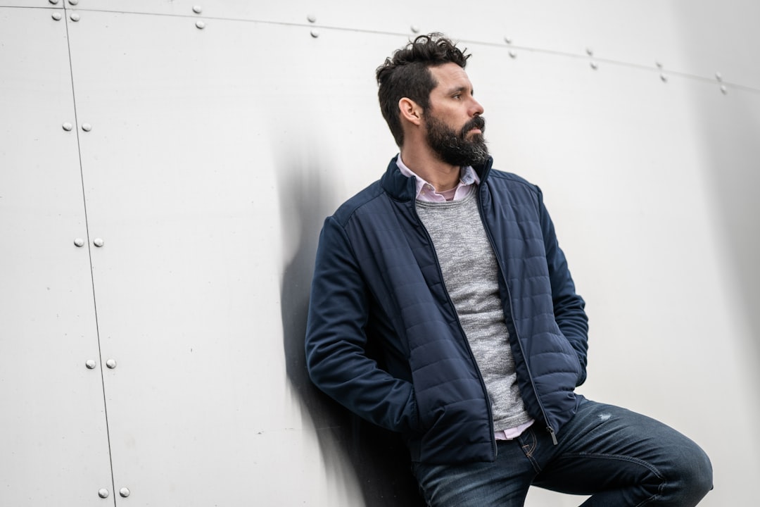 man in blue zip up jacket and blue denim jeans sitting on white concrete wall