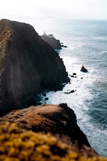 A rugged coastline with dark cliffs meeting the ocean. A small lighthouse sits atop the cliff, surrounded by a few buildings. Waves crash against the rocks, creating white foam along the shoreline. The foreground contains some blurred vegetation, suggesting it is captured from higher ground.