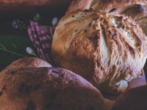 Close-up of freshly baked bread loaves resting on a patterned cloth, with a rustic and artisanal appearance. The bread has a golden-brown crust and is surrounded by soft lighting and subtle background elements, including some green leaves and out-of-focus details.