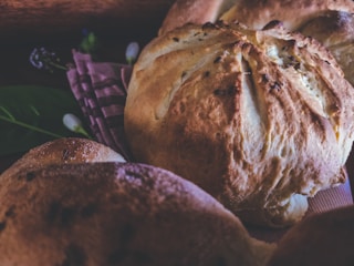 Close-up of freshly baked bread loaves resting on a patterned cloth, with a rustic and artisanal appearance. The bread has a golden-brown crust and is surrounded by soft lighting and subtle background elements, including some green leaves and out-of-focus details.