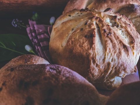 Close-up of freshly baked bread loaves resting on a patterned cloth, with a rustic and artisanal appearance. The bread has a golden-brown crust and is surrounded by soft lighting and subtle background elements, including some green leaves and out-of-focus details.