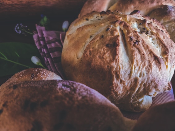 Close-up of freshly baked bread loaves resting on a patterned cloth, with a rustic and artisanal appearance. The bread has a golden-brown crust and is surrounded by soft lighting and subtle background elements, including some green leaves and out-of-focus details.