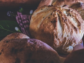 Close-up of freshly baked bread loaves resting on a patterned cloth, with a rustic and artisanal appearance. The bread has a golden-brown crust and is surrounded by soft lighting and subtle background elements, including some green leaves and out-of-focus details.