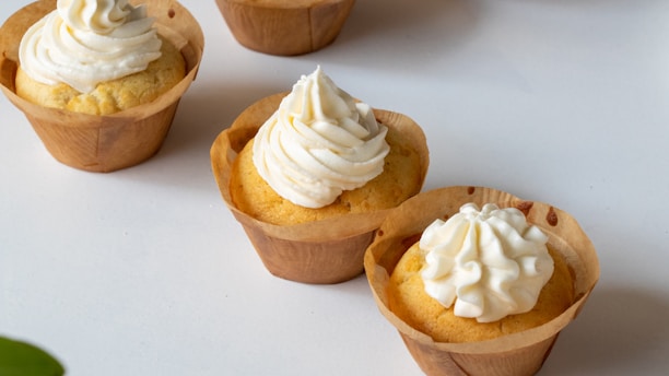 Several cupcakes with a light yellow cake base and topped with swirls of creamy white frosting are placed on a clean white surface. The cupcakes are encased in brown paper liners.