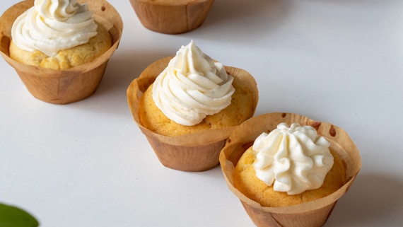 Several cupcakes with a light yellow cake base and topped with swirls of creamy white frosting are placed on a clean white surface. The cupcakes are encased in brown paper liners.