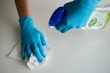 Hands wearing gloves wiping a wooden table with a soft cloth and cleaning spray.