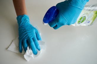 A person wearing blue gloves is cleaning a surface using a paper towel and a spray bottle of cleaning solution.