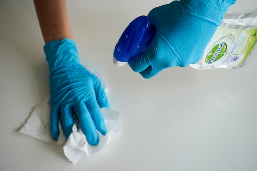 A person wearing blue gloves is cleaning a surface using a paper towel and a spray bottle of cleaning solution.