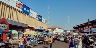A bustling street market scene with numerous people walking and interacting. Cars are parked and moving through the street, creating a vibrant, dynamic atmosphere. Large billboards advertising mobile phones adorn the sides of a tall building. Many colorful umbrellas cover various stalls lined along the street, with vendors selling their goods.
