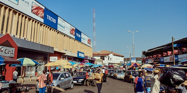 A bustling street market scene with numerous people walking and interacting. Cars are parked and moving through the street, creating a vibrant, dynamic atmosphere. Large billboards advertising mobile phones adorn the sides of a tall building. Many colorful umbrellas cover various stalls lined along the street, with vendors selling their goods.