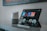 A tablet is placed on a table with its stand, displaying a music streaming interface with multiple album covers and titles. Beside it is a smart speaker with a minimalist design, featuring a white top and gray base. The setting is indoors, possibly a home or office space.