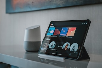 A tablet is placed on a table with its stand, displaying a music streaming interface with multiple album covers and titles. Beside it is a smart speaker with a minimalist design, featuring a white top and gray base. The setting is indoors, possibly a home or office space.