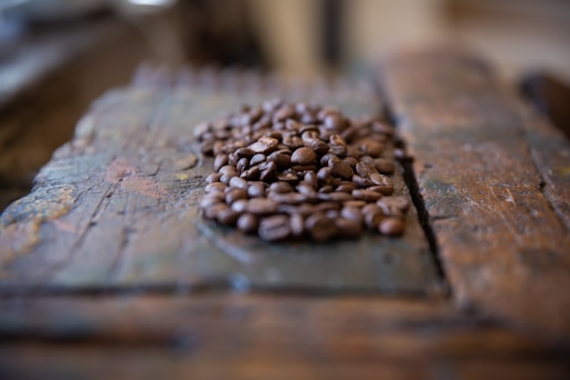 A pile of coffee beans lies on a rustic, wooden surface. The focus is on the beans, which are rich in brown tones, while the background is artistically blurred, enhancing the texture and color contrasts.
