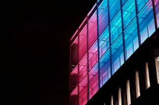 A modern building facade is illuminated with a striking pattern of colorful lights. The structure features a grid-like pattern with a gradient of pink to blue lights transitioning across its surface, creating a vibrant and futuristic appearance against a dark background.