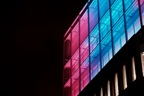 A modern building facade is illuminated with a striking pattern of colorful lights. The structure features a grid-like pattern with a gradient of pink to blue lights transitioning across its surface, creating a vibrant and futuristic appearance against a dark background.