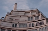 A modern, multi-story apartment building features a series of curved balconies with railings. The facade is smooth with large windows and roller shutters covering some of them. The sky above is clear with a soft blue hue.