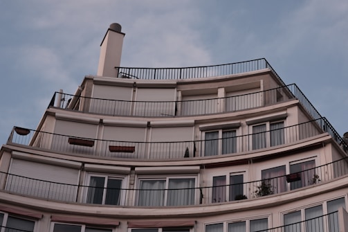 A modern, multi-story apartment building features a series of curved balconies with railings. The facade is smooth with large windows and roller shutters covering some of them. The sky above is clear with a soft blue hue.