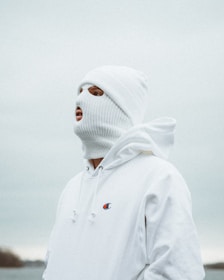 A person wearing a white knitted ski mask and a white hoodie stands in an outdoor setting under a cloudy sky. The logo on the hoodie is visible, and the colors are muted and cool, with a neutral background.