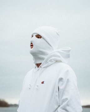 A person wearing a white knitted ski mask and a white hoodie stands in an outdoor setting under a cloudy sky. The logo on the hoodie is visible, and the colors are muted and cool, with a neutral background.