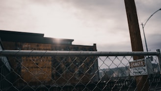 A chain-link fence with a sign labeled 'Danger' and 'Power' is situated in the foreground. Beyond the fence, there is a blurred view of an industrial building under a cloudy sky. The lighting is subdued, suggesting a late afternoon or overcast day.
