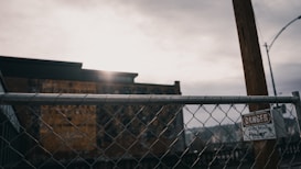A chain-link fence with a sign labeled 'Danger' and 'Power' is situated in the foreground. Beyond the fence, there is a blurred view of an industrial building under a cloudy sky. The lighting is subdued, suggesting a late afternoon or overcast day.