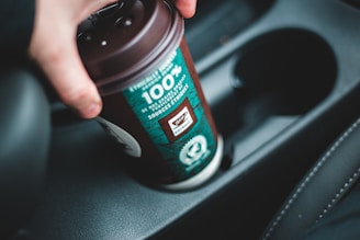 A hand is placing a brown and teal coffee cup into a car cup holder. The cup features a label that highlights ethically sourced ingredients.