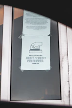 A printed notice taped to a window, indicating a preference for debit or credit card payments over cash. The sign includes a graphic of a hand holding a card. Another message is posted above, possibly policy or informational text.