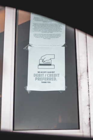 A printed notice taped to a window, indicating a preference for debit or credit card payments over cash. The sign includes a graphic of a hand holding a card. Another message is posted above, possibly policy or informational text.