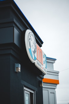 The image captures a close-up view of a drive-thru restaurant building featuring a prominent sign with a logo and text. The sign includes a stylized illustration of a person's face, along with the letters 'MB' and text that reads 'Drive Thru'. The building has a dark facade with modern architectural elements, and there is an overcast sky in the background.