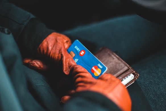 A person wearing brown leather gloves holds a wallet and a blue debit card. The debit card has contactless payment symbols and recognizable brand logos on it. The background is a dark fabric upholstery.