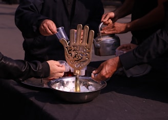 Several people are participating in an activity involving a large bowl filled with sugar cubes. A brass ornament shaped like a hand with intricate inscriptions is placed in the bowl. One person is holding a kettle, while others are pouring liquid into cups.
