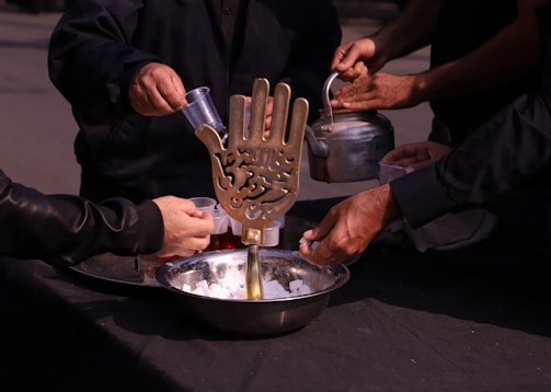 Several people are participating in an activity involving a large bowl filled with sugar cubes. A brass ornament shaped like a hand with intricate inscriptions is placed in the bowl. One person is holding a kettle, while others are pouring liquid into cups.