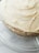 Close-up view of a cake with thick, creamy white frosting, placed on a glass plate. The layers of the cake are partially visible through the frosting, giving it a homemade and rustic appearance.