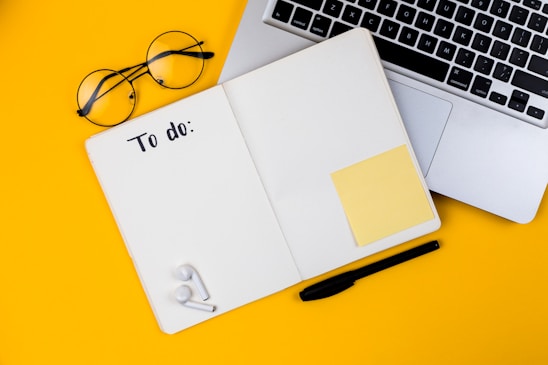 An open notebook with 'To do:' written at the top of a blank page, resting on a yellow surface. Nearby are a pair of eyeglasses, a pen, and wireless earphones. Partially visible is a laptop with a silver metallic finish.