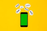 A smartphone with a green screen lies on a bright yellow background, surrounded by speech bubbles containing words like 'Help!', 'Covid-19', 'Oh no!', 'death', and 'Medic'. The arrangement suggests an alarming or urgent context.